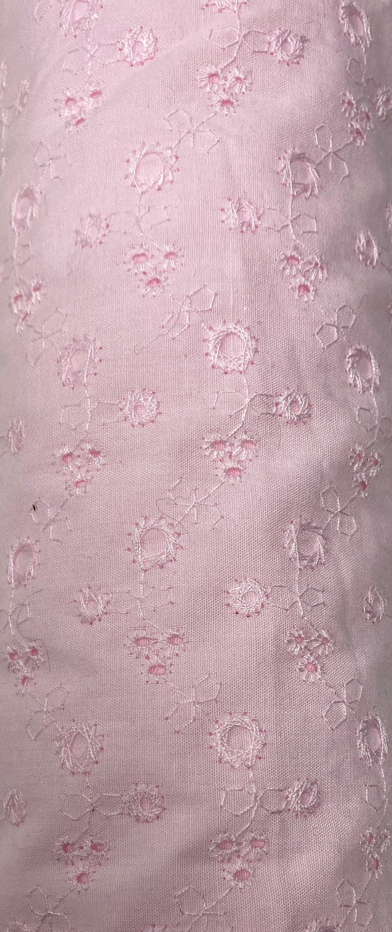 May include: A close-up of a pale pink fabric with a delicate floral pattern. The fabric has a subtle sheen and a soft, airy texture.