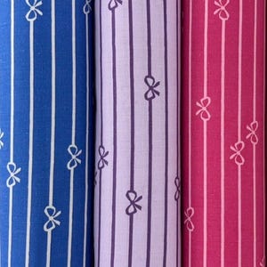 May include: Close-up of three rolls of fabric in blue, lavender, and pink. Each roll features vertical stripes with a repeating bow pattern. The blue fabric has white stripes, the lavender has purple stripes, and the pink has white stripes.