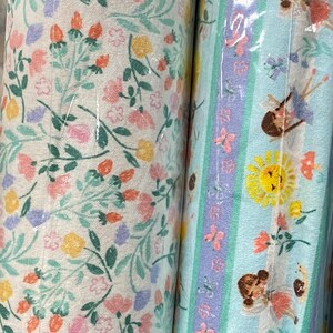 May include: Two fabric rolls with floral and fairy designs. The first roll features a white background with a small floral pattern in shades of pink, yellow, and green. The second roll features a light blue background with a striped pattern in shades of purple, green, and white. The pattern includes fairies, butterflies, and sunflowers.