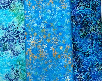 May include: Three pieces of blue and green batik fabric with floral patterns. The fabric on the left has a swirling pattern, the fabric in the middle has a pattern of flowers and stars, and the fabric on the right has a more traditional floral pattern.