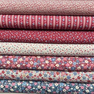 May include: A stack of seven folded fabric bolts featuring various floral and geometric patterns in shades of red, pink, blue, and white. The designs include small leaf prints, vertical stripes with tiny flowers, and dense floral motifs.