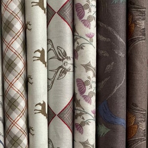May include: Five fabric swatches with different patterns. The first swatch is a plaid pattern in shades of brown, green, and white. The second swatch features a beige background with brown deer silhouettes. The third swatch has a beige background with a brown deer silhouette and a diamond pattern in shades of brown and red. The fourth swatch has a beige background with a purple thistle pattern. The fifth swatch is a brown fabric with a dark blue bird silhouette and a green and brown leaf pattern.