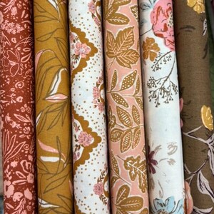 May include: Close-up of several rolls of fabric with floral and leaf patterns. The fabrics feature a variety of colours, including rust, mustard yellow, pink, and white. The designs include detailed botanical illustrations.