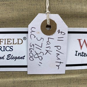 May include: Close-up of tan fabric with a woven texture. A white tag is attached with a string, displaying handwritten text including "Lark 57158" and "uc 5600". The tag also includes the price per metre.