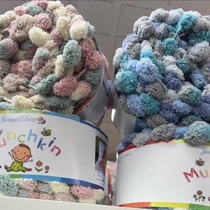 May include: Two spools of Munchkin yarn in pastel shades. One has pink, cream, and mint green pom-poms, the other blue, grey, and teal. The label reads "Munchkin" with a baby graphic. Ideal for knitting and crochet projects.