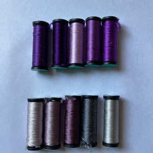 May include: A collection of spools of thread in various shades of purple, grey, and silver. The spools are arranged in two rows, with the purple spools on top and the grey and silver spools on the bottom.