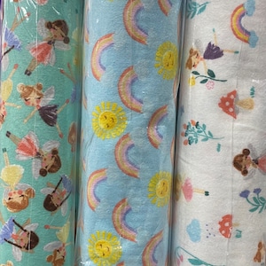 May include: Three rolls of fabric with different patterns. The first roll has a light blue background with rainbows and yellow suns. The second roll has a white background with rainbows, flowers, and mushrooms. The third roll has a light green background with fairies and flowers.
