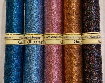 Può includere: Sei bobine di filo metallico in vari colori, tra cui blu, blu-verde, rosa, oro e nero. Le bobine sono etichettate con il nome del marchio "Gütermann" e la composizione del filo, tra cui poliestere e poliammide.