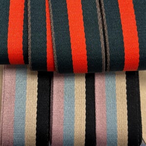 May include: Close-up of two different striped ribbons. The top ribbon has alternating vertical stripes of dark teal and bright orange. The bottom ribbon has stripes of black, light pink, light blue, and beige. A ruler is at the bottom.