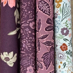 May include: Close-up of four fabric rolls with floral and leaf patterns. The colour palette includes shades of purple, pink, and white. The designs feature various flower and leaf motifs, suitable for sewing and crafting projects.