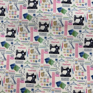 May include: Cream-coloured fabric featuring a repeating pattern of sewing machines, thread spools, scissors, and other haberdashery items. The words "100% Handmade" and "Handmade" are printed on the fabric. The design includes pink, blue, and green elements.