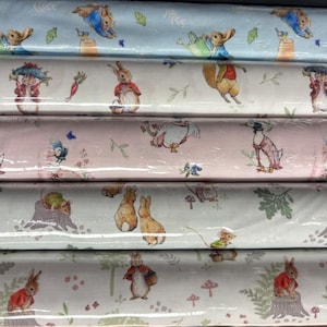May include: Five stacked bolts of fabric featuring Peter Rabbit illustrations. The patterns include rabbits in blue jackets, carrots, ducks, and mice on light blue, white, pink, and pale green backgrounds with small leaves, flowers, and mushrooms.