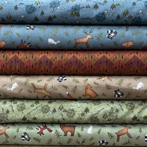 May include: A pile of fabric rolls featuring woodland animal and landscape designs. Patterns include deer, foxes, bears, and badgers, alongside compasses, trees, and mountains. The colour palette includes blues, greens, browns, and tans, giving a rustic feel.