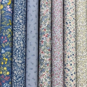 May include: Several rolls of patterned fabric, showcasing floral designs in blue, pink, and cream. The fabrics feature small, detailed flower and leaf patterns, ideal for sewing and craft projects. The rolls are tightly wound.