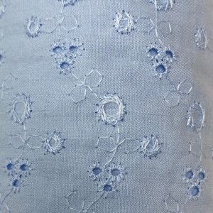 May include: A close-up of a light blue fabric with a delicate white eyelet pattern. The fabric has a subtle texture and a soft, airy feel.