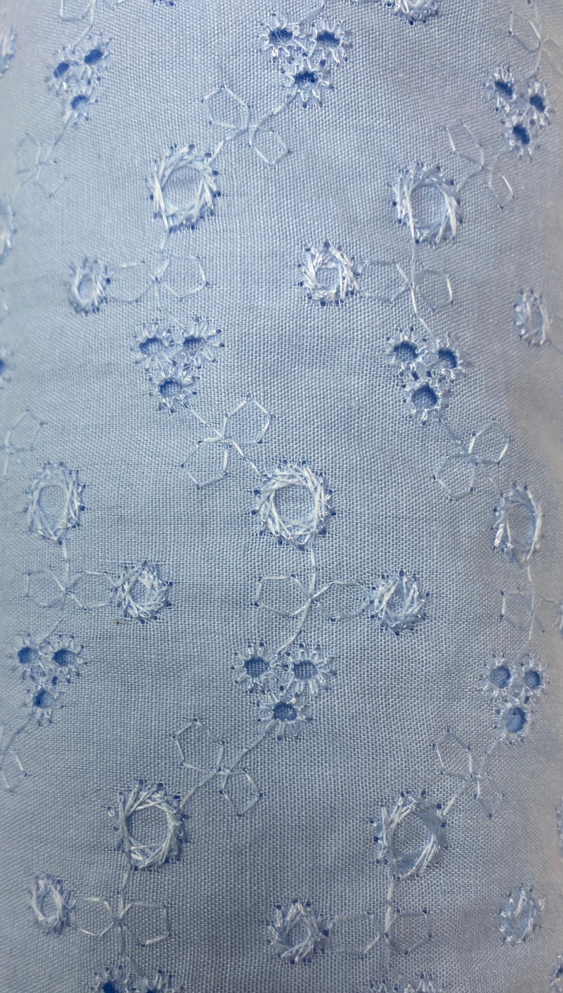 May include: A close-up of a light blue fabric with a delicate white eyelet pattern. The fabric has a subtle texture and a soft, airy feel.