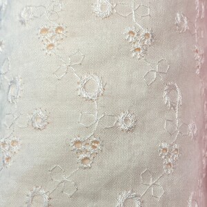 May include: White fabric with a delicate embroidered floral pattern. The fabric has a subtle texture and a soft, airy feel.