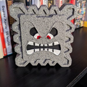 May include: A gray, textured, block-shaped figure with a cartoon face. The figure has black eyes, red pupils, a black mouth, and white teeth. The figure is sitting on a black shelf with video game cases behind it.