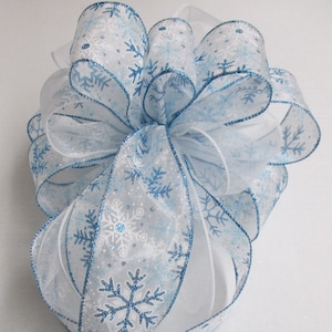 May include: A decorative bow made of white and light blue snowflake patterned ribbon. The ribbon has a blue glitter trim and is tied into a large bow. The bow is suitable for winter or holiday decorations.