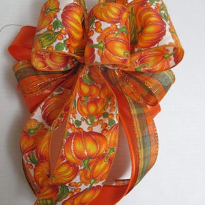 May include: A decorative bow crafted from multiple ribbons, featuring a white background with a vibrant orange and yellow pumpkin print, interspersed with green leaves. The design includes solid orange satin ribbons and plaid ribbons.
