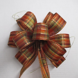 May include: A decorative bow made from wired ribbon featuring a plaid pattern in shades of orange, brown, and green. The ribbon has a metallic, shimmering finish with thin gold-toned wire edges and includes two thin wire ties extending from the center.