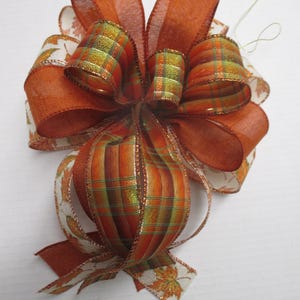 May include: A decorative bow crafted from multiple ribbons, featuring solid burnt orange, a plaid pattern in orange, green, and gold, and a cream-colored ribbon with a printed pattern of autumn leaves in shades of orange, yellow, and brown.