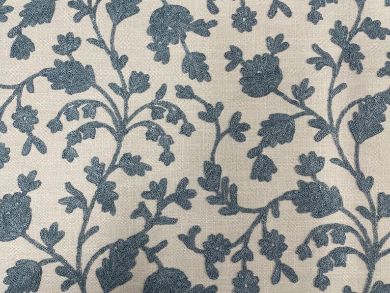 May include: A cream-colored fabric with a blue embroidered floral pattern. The design features delicate branches with small flowers and leaves.