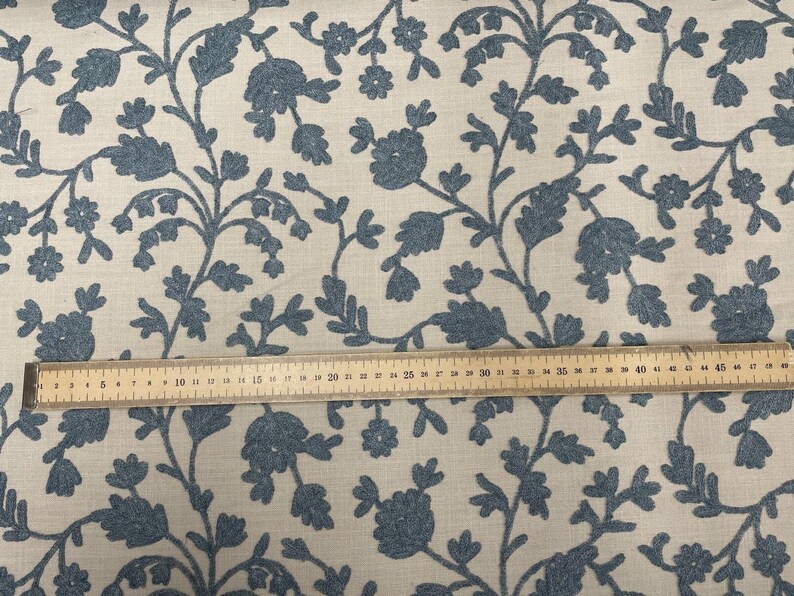 May include: Blue floral embroidery on a cream-colored fabric. A wooden ruler is placed on the fabric for scale.