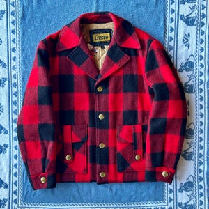 May include: A red and black plaid jacket with gold buttons and two front pockets. The jacket has a collar and the label reads "Cresco". The jacket is made of a thick, warm material.