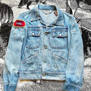 May include: Light blue denim jacket with a button-up front, two chest pockets, and two side pockets. The jacket features patches on the sleeves, one with a car and the other with an eagle. The jacket has a classic collar and buttoned cuffs.