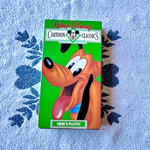 May include: A Walt Disney Cartoon Classics VHS tape featuring Pluto. The box is green with the Disney logo and Pluto's image. The text "HERE'S PLUTO!" is printed on the bottom of the box.