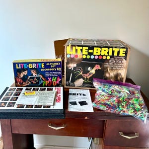 May include: A vintage Lite-Brite toy set with a black and white box, a clear plastic bag of colorful pegs, and a black and white instruction booklet. The box has the text "Lite-Brite Alphabet Set accessory kit" and "SPELL OUT WORDS AND SENTENCES WITH YOUR LIGHT UP LETTERS".
