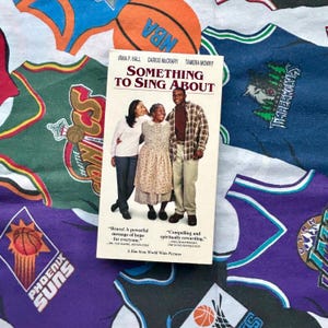 May include: A VHS tape titled "Something to Sing About" featuring Irma P. Hall, Darius McCrary, and Tamera Mowry. The cover shows three people and text. The tape rests on a colorful background of sports-themed fabric with basketball and team logos.
