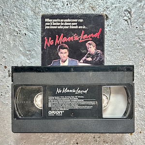 May include: A black VHS tape with the title "No Man's Land" printed on the label. The tape is sitting on a grey concrete surface. The movie is rated R and has a running time of 107 minutes.