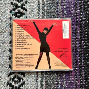 May include: A red and black CD case with a photo of a woman wearing a black dress and holding her arms up. The CD case is for the album "Another Sad Love Song" by Toni Braxton. The track list is printed on the back of the case.