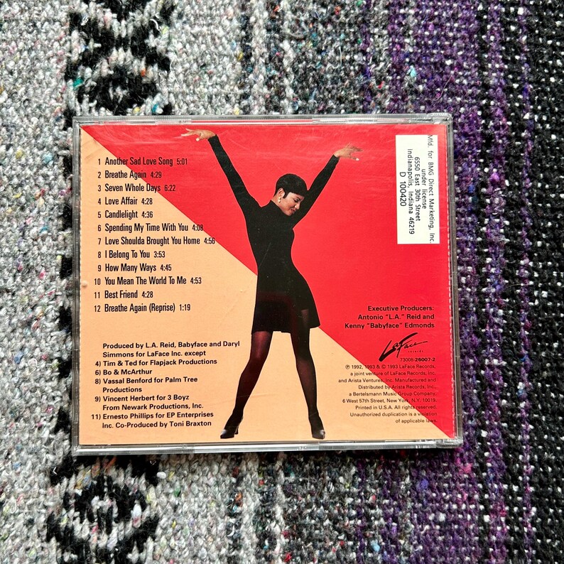 May include: A red and black CD case with a photo of a woman wearing a black dress and holding her arms up. The CD case is for the album "Another Sad Love Song" by Toni Braxton. The track list is printed on the back of the case.