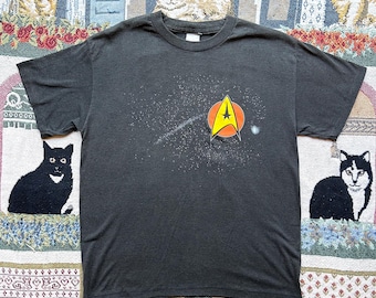 May include: Dark gray t-shirt featuring a Star Trek emblem with a yellow star and a red circle. The shirt has a crew neck and short sleeves. The graphic includes a white streak and small white dots.