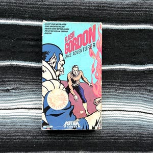 May include: A vintage Flash Gordon Space Adventurer VHS tape. The cover features a comic book style illustration with the title in red and a central figure in a brown shirt and blue pants. The background is blue and pink.