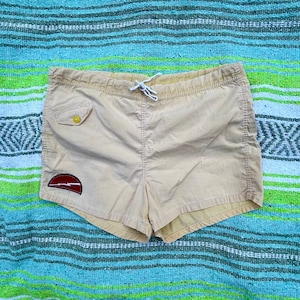 May include: Beige swim trunks with a red and yellow patch on the left leg. The trunks have a drawstring waist and a side pocket.