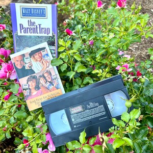 Puede incluir: Una cinta VHS y una caja de DVD de la película "La trampa de los padres" con una foto de los actores en la parte delantera. La caja está encima de un arbusto con flores rosas.