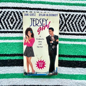 May include: A paperback book titled "Jersey Girl" featuring Jami Gertz and Dylan McDermott. The cover art depicts a woman in a pink top and polka dot skirt, and a man in a suit. The book's tagline is "A Cinderella story with big hair."