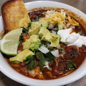 May include: A bowl of chili topped with shredded cheese, sour cream, avocado, lime, and cilantro.