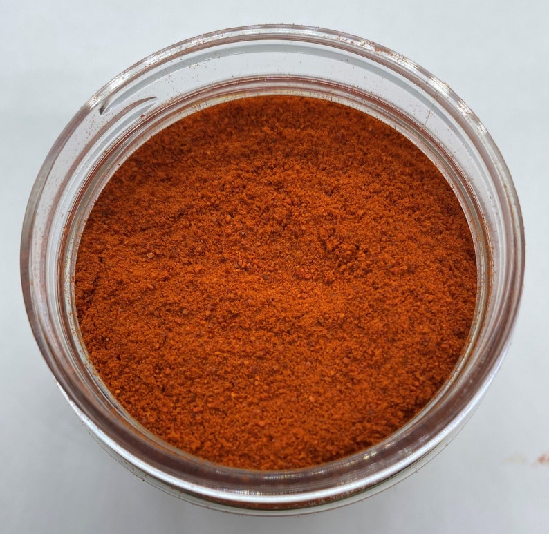 May include: A clear glass jar filled with a fine, bright red powder. The powder appears to be a spice or seasoning, with a texture similar to paprika or chili powder. The jar is open, revealing the contents within.