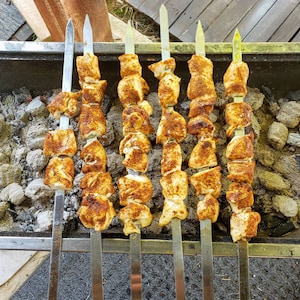 May include: Close-up of six skewers of chicken, each with multiple pieces of chicken, on a charcoal grill. The chicken is seasoned and cooked, with a golden brown color. The skewers are arranged in a row on the grill, with the tips of the skewers visible at the top and bottom of the image.