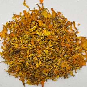 May include: A pile of dried marigold flower petals in various shades of vibrant orange and golden yellow. The petals are thin, curled, and textured, scattered in a loose, circular arrangement against a plain, neutral white background.