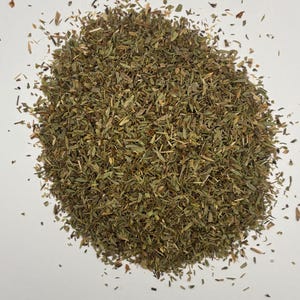 May include: A pile of dried, chopped green herbs, possibly tarragon or thyme, against a white background. The herbs are a mix of light and dark greens, with some small, brown pieces scattered throughout. The image is a close-up, showing the texture of the herbs.