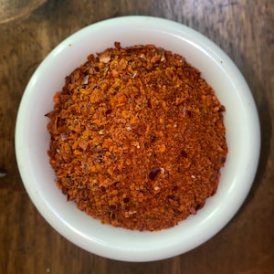 May include: A white bowl filled with a generous amount of crushed red pepper flakes. The flakes are a vibrant red-orange color and appear to be finely ground.