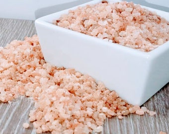 May include: A white ceramic bowl filled with pink Himalayan salt crystals. The salt is spilling out of the bowl onto a wooden surface.