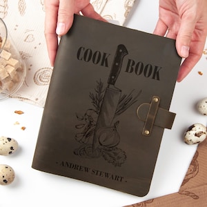Puede incluir: Un libro de cocina de cuero marrón oscuro con las palabras "COOK BOOK" y una ilustración de cuchillo. El libro de cocina tiene un cierre de correa y el nombre "- ANDREW STEWART -" impreso en la parte inferior. El libro es sostenido por dos manos.