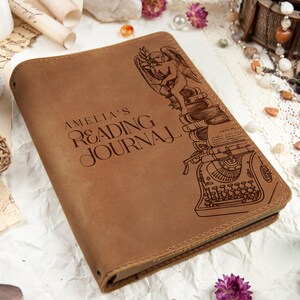 Puede incluir: Un diario de cuero marr&oacute;n con el texto "AMELIA'S READING JOURNAL" en relieve en la portada. Una ilustraci&oacute;n de un &aacute;ngel, libros y una m&aacute;quina de escribir est&aacute; en el lado derecho. El diario est&aacute; cerrado y tiene un borde cosido.